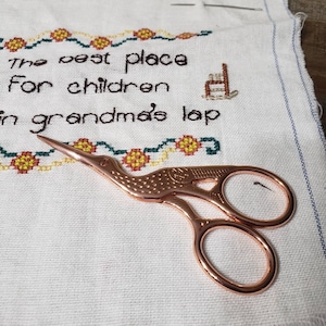 May include: A pair of rose gold embroidery scissors with a bird design. The scissors are lying on a white fabric with cross-stitch embroidery that reads "The best place for children in grandma's lap".