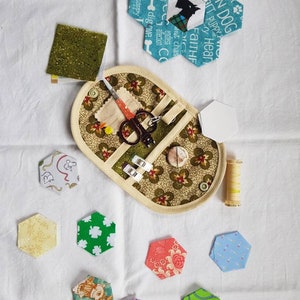 Peut inclure: Une trousse de couture en tissu beige avec un motif floral et une fermeture à glissière. La trousse contient une paire de ciseaux, une bobine de fil et quelques épingles à coudre. La trousse est entourée de divers hexagones en tissu colorés.