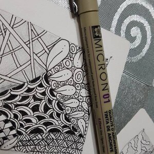Puede incluir: Primer plano de un bol&iacute;grafo Sakura Micron 01 con una l&iacute;nea de 0,25 mm. El bol&iacute;grafo es beige con detalles negros y presenta el texto "QUALIT&Eacute; D'ARCHIVAGE TINTA DE ARCHIVE" en morado. El bol&iacute;grafo est&aacute; sobre una superficie blanca con un dise&ntilde;o de garabatos en blanco y negro.