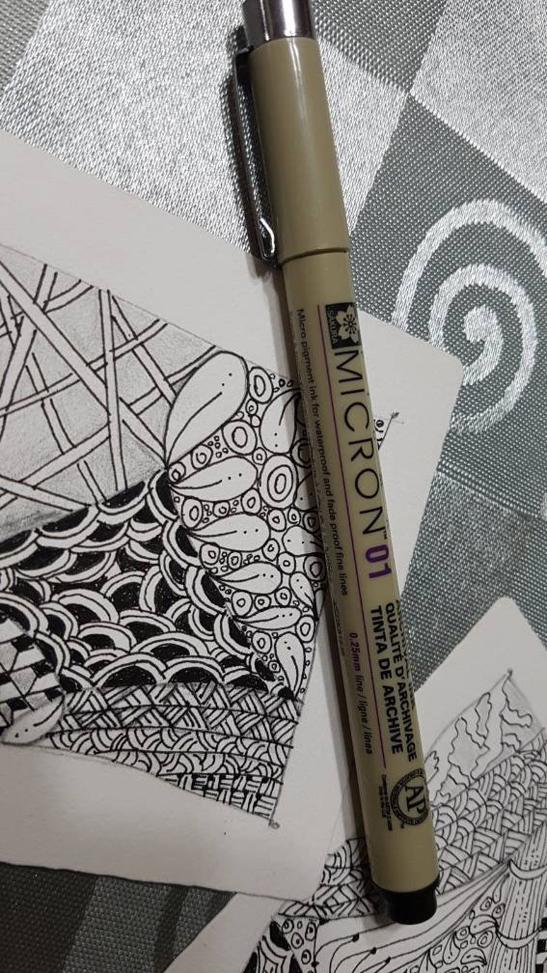 Puede incluir: Primer plano de un bol&iacute;grafo Sakura Micron 01 con una l&iacute;nea de 0,25 mm. El bol&iacute;grafo es beige con detalles negros y presenta el texto "QUALIT&Eacute; D'ARCHIVAGE TINTA DE ARCHIVE" en morado. El bol&iacute;grafo est&aacute; sobre una superficie blanca con un dise&ntilde;o de garabatos en blanco y negro.