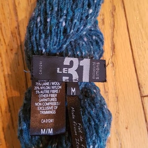 Pode incluir: Um novelo de fio azul-esverdeado com manchas brancas. O r&oacute;tulo do fio diz "Le 31" e "exclusivit&eacute; Simons" e indica que o fio &eacute; feito de fio italiano. O r&oacute;tulo tamb&eacute;m lista a composi&ccedil;&atilde;o do fio como 75% l&atilde;, 20% nylon e 5% outra fibra. O r&oacute;tulo indica que o fio &eacute; do tamanho M/M.