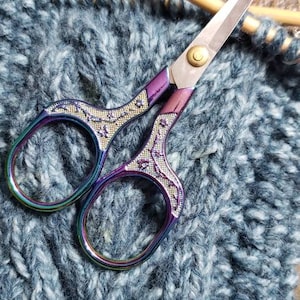 Peut inclure: Ciseaux décoratifs avec une poignée et des lames aux couleurs de l'arc-en-ciel, posés sur un tissu tricoté bleu. Les ciseaux ont un motif violet et argenté sur les poignées et une vis dorée.