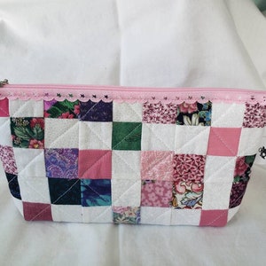 May include: A pink and white patchwork zippered pouch with a floral print. The pouch is quilted and has a white background with pink and purple squares.
