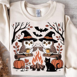 May include: Cream-colored sweatshirt featuring a Halloween-themed graphic. Two owls wearing witch hats sit on a log, holding mugs and marshmallows. Pumpkins, a black cat, bats, and a campfire complete the design.
