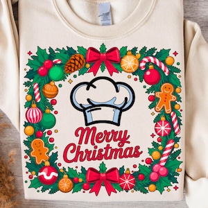 May include: A cream-colored sweatshirt with a festive Christmas design. The graphic features a chef's hat, a wreath with ornaments, gingerbread men, and the words "Merry Christmas" in red. The wreath includes red bows, candy canes, and pinecones.