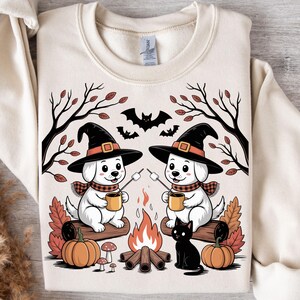 May include: Cream-colored sweatshirt featuring a Halloween-themed illustration. Two white puppies wearing witch hats and scarves are roasting marshmallows over a campfire. Pumpkins, mushrooms, a black cat, and bats complete the fall scene.