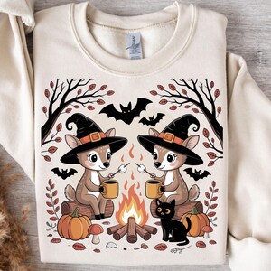 May include: Beige sweatshirt featuring a Halloween-themed graphic. The design includes two deer wearing witch hats, roasting marshmallows over a campfire, with pumpkins, bats, and a black cat. The sweatshirt has a crew neck and long sleeves.