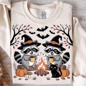 May include: Cream-colored sweatshirt featuring a Halloween-themed design. Two cartoon raccoons wearing witch hats sit by a campfire, roasting marshmallows. A black cat, pumpkins, bats, and fall foliage complete the festive illustration.