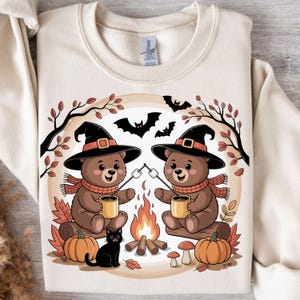 May include: Cream-colored sweatshirt featuring a Halloween-themed graphic. Two brown bears wearing witch hats and scarves sit by a campfire, roasting marshmallows. A black cat, pumpkins, and bats complete the autumn scene.