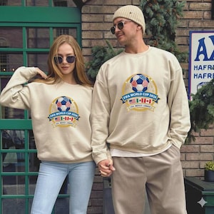 May include: Two people wearing beige sweatshirts with a FIFA World Cup 2026 design. The design includes a soccer ball, the text "FIFA WORLD CUP 2026", and the flags of the USA, Mexico, and Canada. The sweatshirts are long-sleeved.