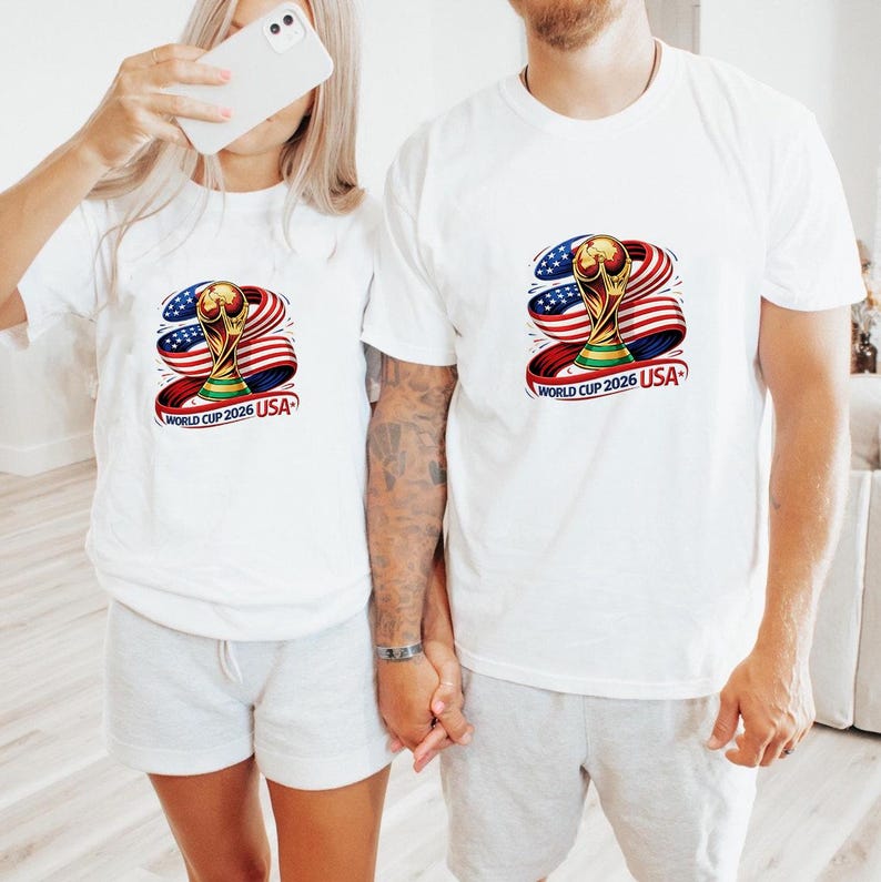 May include: White t-shirts featuring a colorful graphic of the World Cup trophy, the American flag, and the text "WORLD CUP 2026 USA". The shirts are worn by two people, one holding a phone.
