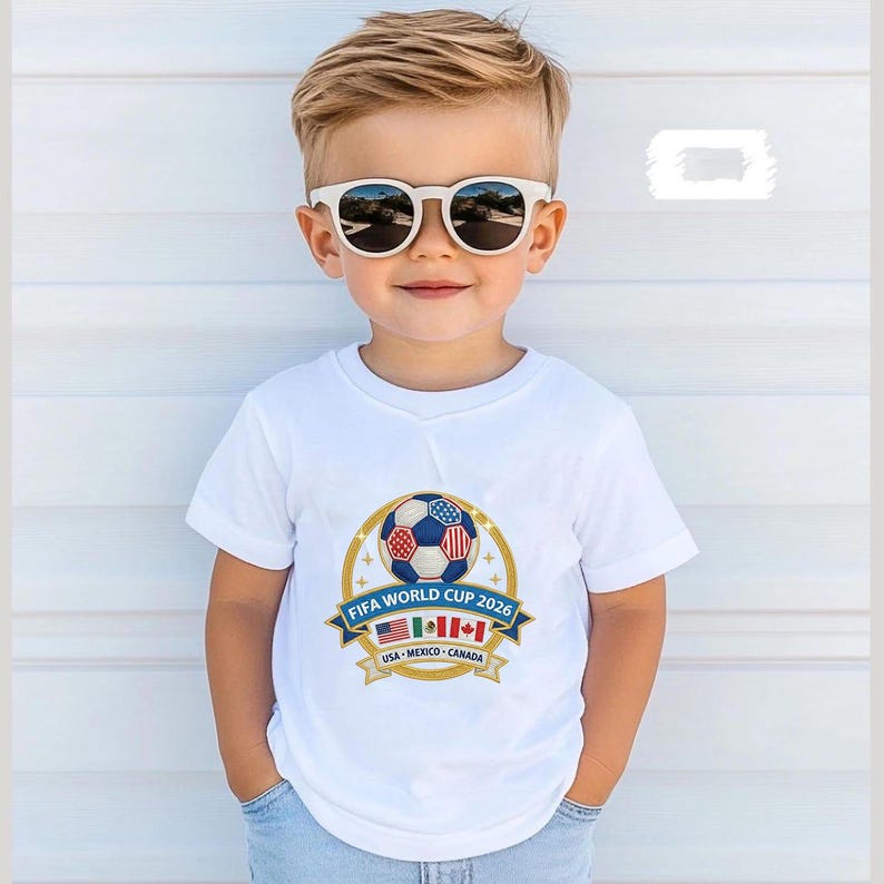 May include: A young child wearing a white t-shirt with a soccer ball graphic and the text "FIFA WORLD CUP 2026". The graphic includes the flags of the USA, Mexico, and Canada. The child is also wearing white sunglasses.