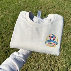 May include: Cream-colored sweatshirt with a crew neck. The front features a colorful emblem with a soccer ball and the text "FIFA WORLD CUP 2026" with the flags of USA, Mexico, and Canada. The sweatshirt is folded.