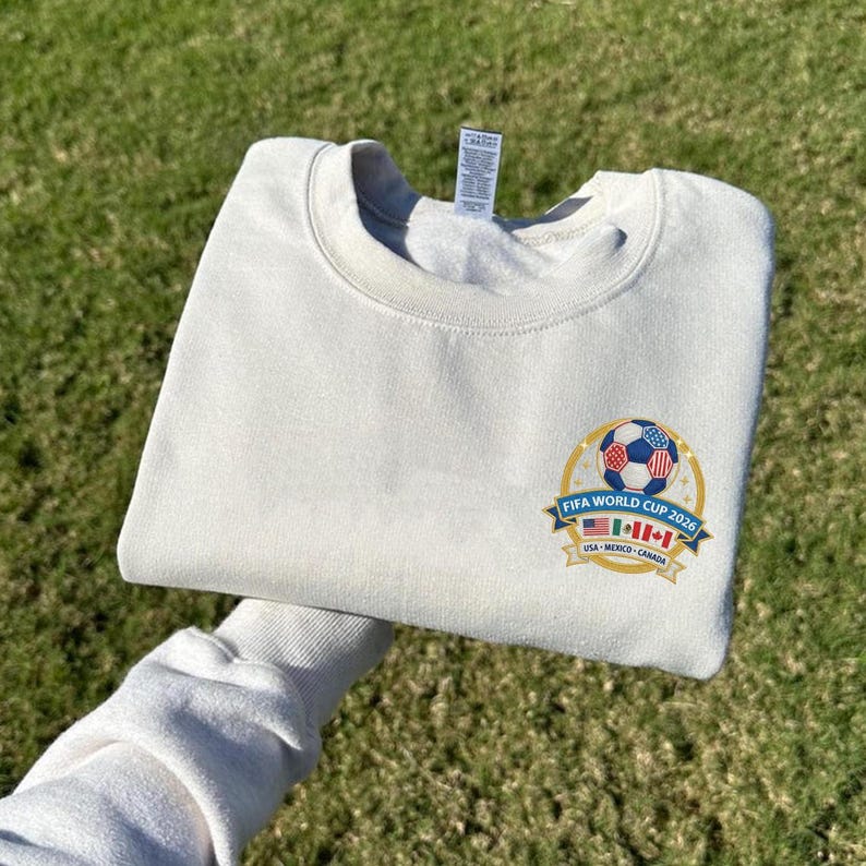 May include: Cream-colored sweatshirt with a crew neck. The front features a colorful emblem with a soccer ball and the text "FIFA WORLD CUP 2026" with the flags of USA, Mexico, and Canada. The sweatshirt is folded.