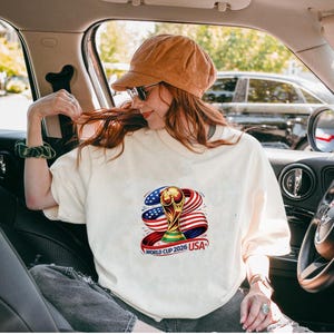 May include: Cream-colored t-shirt featuring a colorful graphic of the World Cup trophy, the American flag, and the text "WORLD CUP 2026 USA". The design includes red, white, blue, and gold colors. The shirt is worn by a person in a car.