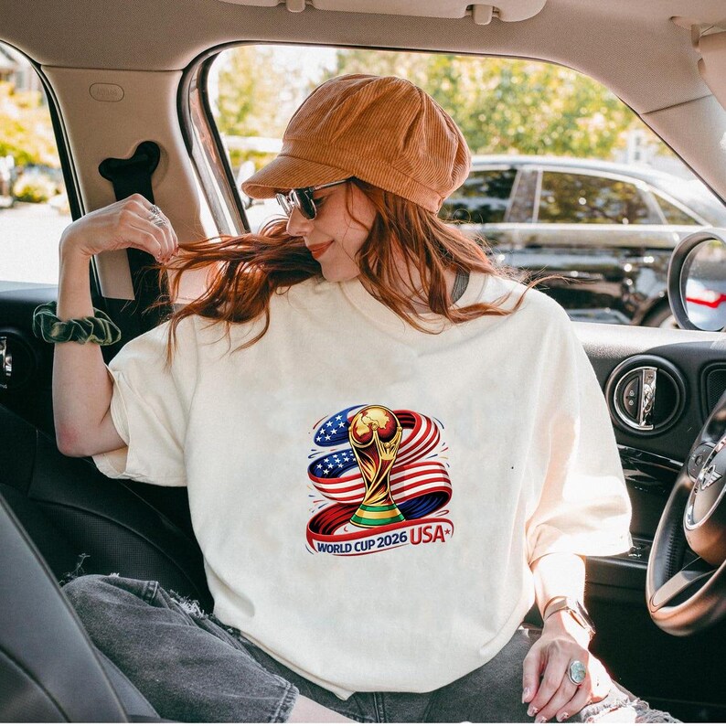 May include: Cream-colored t-shirt featuring a colorful graphic of the World Cup trophy, the American flag, and the text "WORLD CUP 2026 USA". The design includes red, white, blue, and gold colors. The shirt is worn by a person in a car.