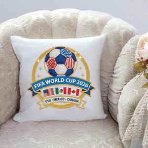 May include: White square pillow with a soccer ball design. The ball is blue, red, and white, surrounded by a gold ring and the text "FIFA WORLD CUP 2026". Below are the flags of USA, Mexico, and Canada.