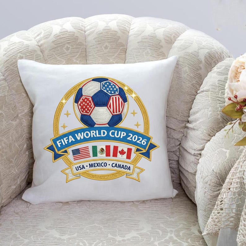 May include: White square pillow with a soccer ball design. The ball is blue, red, and white, surrounded by a gold ring and the text "FIFA WORLD CUP 2026". Below are the flags of USA, Mexico, and Canada.