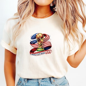 May include: Cream-colored t-shirt featuring a graphic of the World Cup trophy with a red, white, and blue ribbon design. The text "WORLD CUP 2026 USA" is printed below the trophy. The shirt has short sleeves.