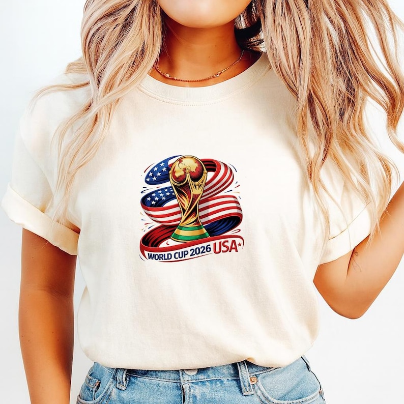 May include: Cream-colored t-shirt featuring a graphic of the World Cup trophy with a red, white, and blue ribbon design. The text "WORLD CUP 2026 USA" is printed below the trophy. The shirt has short sleeves.