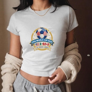May include: A light gray t-shirt featuring a FIFA World Cup 2026 design. The graphic includes a soccer ball with red, white, and blue accents, a blue banner with text, and flags of the USA, Mexico, and Canada.