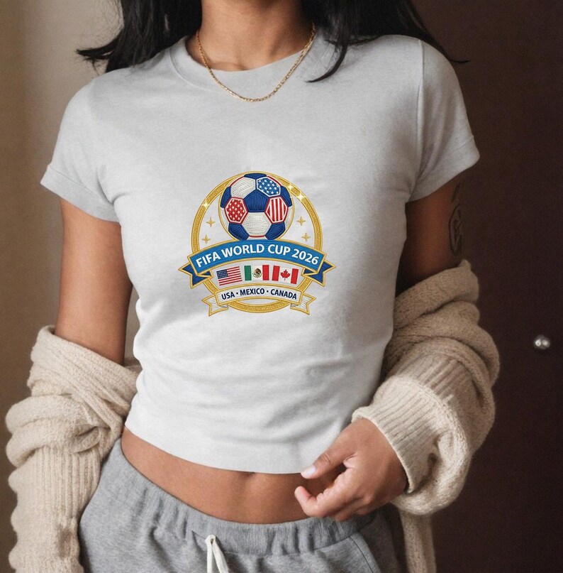 May include: A light gray t-shirt featuring a FIFA World Cup 2026 design. The graphic includes a soccer ball with red, white, and blue accents, a blue banner with text, and flags of the USA, Mexico, and Canada.