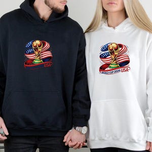 May include: Navy blue and white hooded sweatshirts featuring a graphic of a gold trophy, the American flag, and the text "USA". The sweatshirts have a front pocket and a drawstring hood. The design is centered on the front of the sweatshirts.