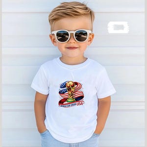May include: A young child wearing a white t-shirt with a graphic of the World Cup trophy, the American flag, and the text "WORLD CUP 2026 USA." The child is also wearing white sunglasses.