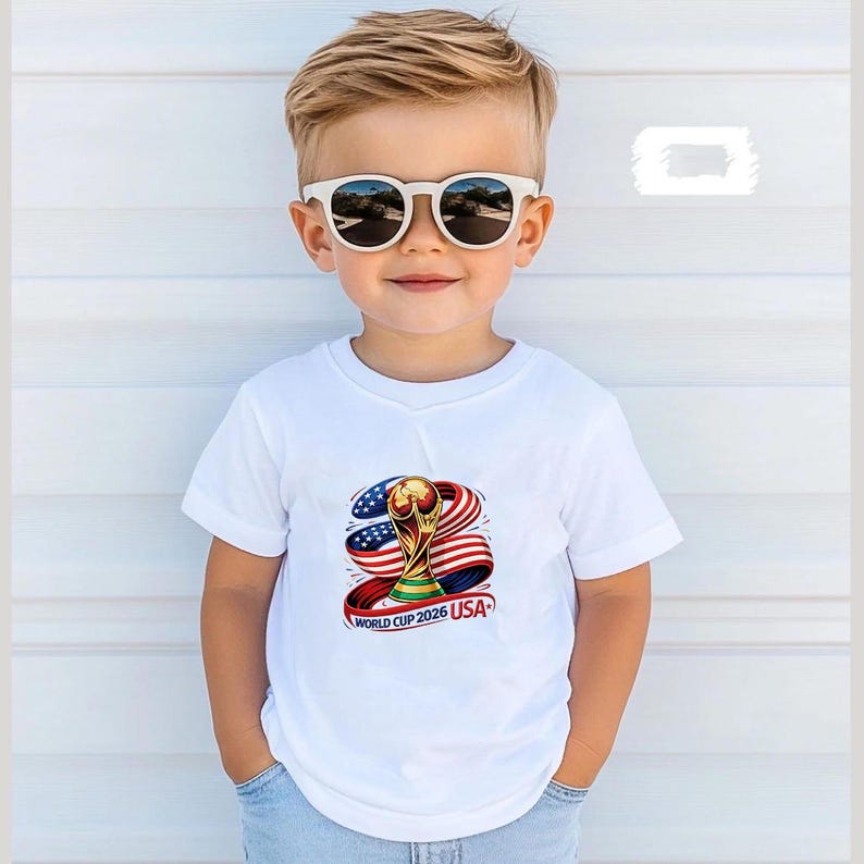May include: A young child wearing a white t-shirt with a graphic of the World Cup trophy, the American flag, and the text "WORLD CUP 2026 USA." The child is also wearing white sunglasses.