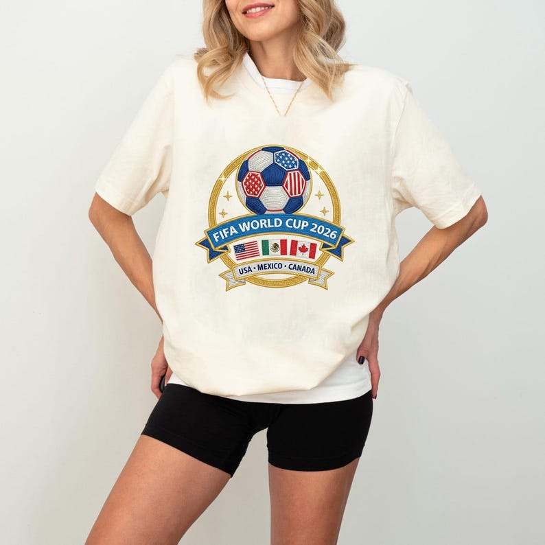 May include: Cream-colored t-shirt featuring a FIFA World Cup 2026 design. The graphic includes a soccer ball with red, white, and blue panels, a banner with the text "FIFA WORLD CUP 2026", and flags of the USA, Mexico, and Canada.