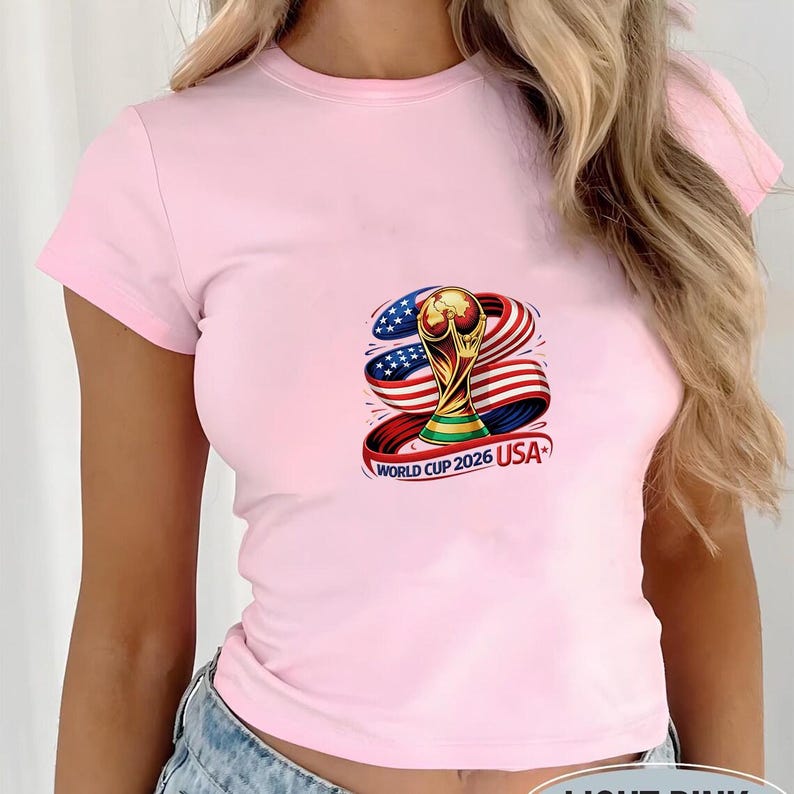 May include: Light pink t-shirt featuring a colorful graphic of the World Cup trophy, the American flag, and the text "WORLD CUP 2026 USA". The shirt has short sleeves and a crew neck.