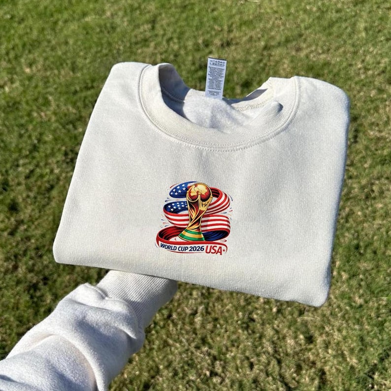 May include: Cream-colored sweatshirt with a crew neck. The front features a colorful graphic of the World Cup trophy, the American flag, and the text "WORLD CUP 2026 USA". The sweatshirt is folded and held up against a green grassy background.