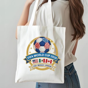 May include: White tote bag featuring a soccer ball design with the text "FIFA WORLD CUP 2026". The design includes the flags of the USA, Mexico, and Canada. The bag has two white handles.