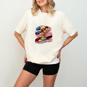 May include: Cream-colored t-shirt featuring a colorful graphic of the World Cup trophy, with red, white, and blue ribbons and the text "WORLD CUP 2026 USA". The shirt has short sleeves and a crew neck.