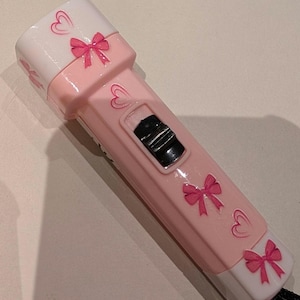 May include: A pink and white flashlight with a black on/off switch. The flashlight is decorated with pink bows and hearts. A black lanyard is attached to the base of the flashlight.