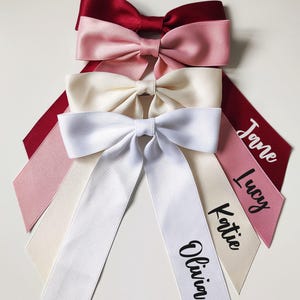 May include: Four satin bows in shades of red, pink, and white. Each bow has long, pointed tails. The tails of three bows are printed with names: Olivia, Katie, and Lucy. The fourth bow has the name Jane.