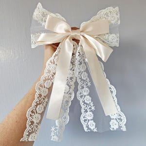 May include: Cream-coloured hair bow with a satin centre and long, flowing ribbons. The bow is trimmed with delicate lace, creating a romantic and elegant accessory. Ideal for special occasions or adding a touch of vintage charm.