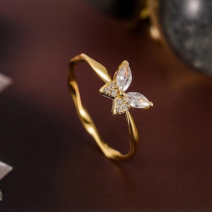 Peut inclure: Une bague dorée ornée d'un motif papillon. Le papillon est composé de deux pierres claires de forme marquise pour les ailes et d'un groupe de petites pierres pour le corps. L'anneau de la bague est torsadé.
