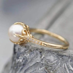 May include: A gold ring featuring a large, round white pearl. The ring has a delicate, ornate design with small, sparkling stones set along the band. The ring is set on a gray surface.