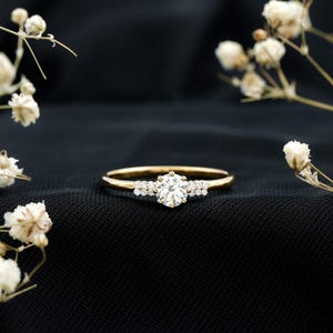 Puede incluir: Un anillo de oro con una gema grande, redonda y transparente en el centro, flanqueada por gemas más pequeñas. El anillo está sobre un fondo negro, con flores blancas en el fondo.