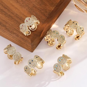 Puede incluir: Anillos dorados con diseño de carita sonriente. Cada anillo tiene una banda que se envuelve alrededor del dedo y está adornado con pequeñas piedras brillantes. Los ojos de las caritas sonrientes están coloreados con piedras rojas, verdes, azules y turquesas.
