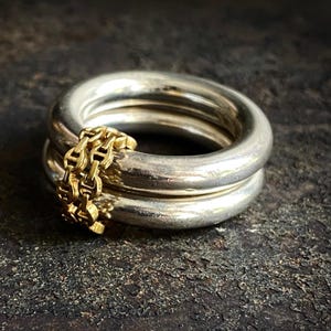 May include: A thick, polished silver band ring featuring a smaller, gold-toned chain-link ring intertwined with it. The silver band has a smooth, rounded profile, while the gold-toned ring consists of small, repeating oval links.