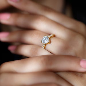 Può includere: Un anello d'oro con un diamante a forma di cuore come pietra centrale. La fascia è impreziosita da diamanti più piccoli. L'anello è indossato su un dito con smalto rosa.