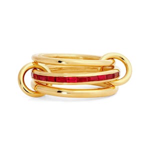 May include: A gold ring featuring three interlocking bands. The central band is inlaid with a continuous row of rectangular red gemstones, while the two outer bands are smooth, polished gold, connected by a small gold loop on the side.