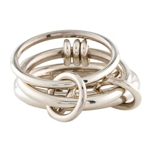 May include: A silver-toned ring composed of multiple interlocking bands. The design features a larger circular band that encircles several smaller, thinner rings, creating a layered, interconnected structure with a smooth, polished metallic finish throughout.