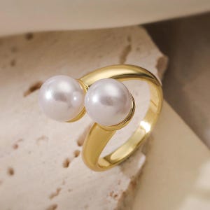 May include: A gold-colored ring featuring two large, round, white pearls. The ring has a modern, open design with a smooth, polished finish. The pearls are set close together, creating a unique and elegant look. The ring is likely made of a gold-toned metal.