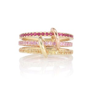 May include: A multi-band ring featuring three interconnected gold bands, each set with a row of small gemstones. The top band contains deep red rubies, the middle band features light pink sapphires, and the bottom band displays yellow sapphires.