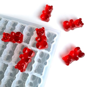 Könnte beinhalten: Nahaufnahme einer transparenten Silikon-Gummibärenform mit mehreren roten Gummibären. Einige Gummibären befinden sich in der Form, andere sind verstreut. Die Form hat mehrere bärenförmige Hohlräume. Die Gummibären sind rot.
