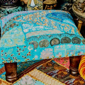 May include: A square, upholstered ottoman with a patchwork design in shades of blue, teal, and gold. The top features various patterns and textures, with dark wooden legs and brass-colored stud detailing along the bottom edge. The ottoman sits on a patterned rug.