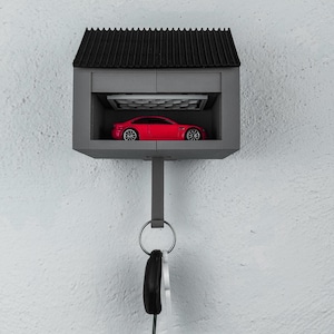 Puede incluir: Un llavero gris en forma de garaje en miniatura con techo negro. Dentro del garaje hay un coche de juguete rojo. Un llavero con llaves cuelga debajo. El llavero está montado en una pared blanca.